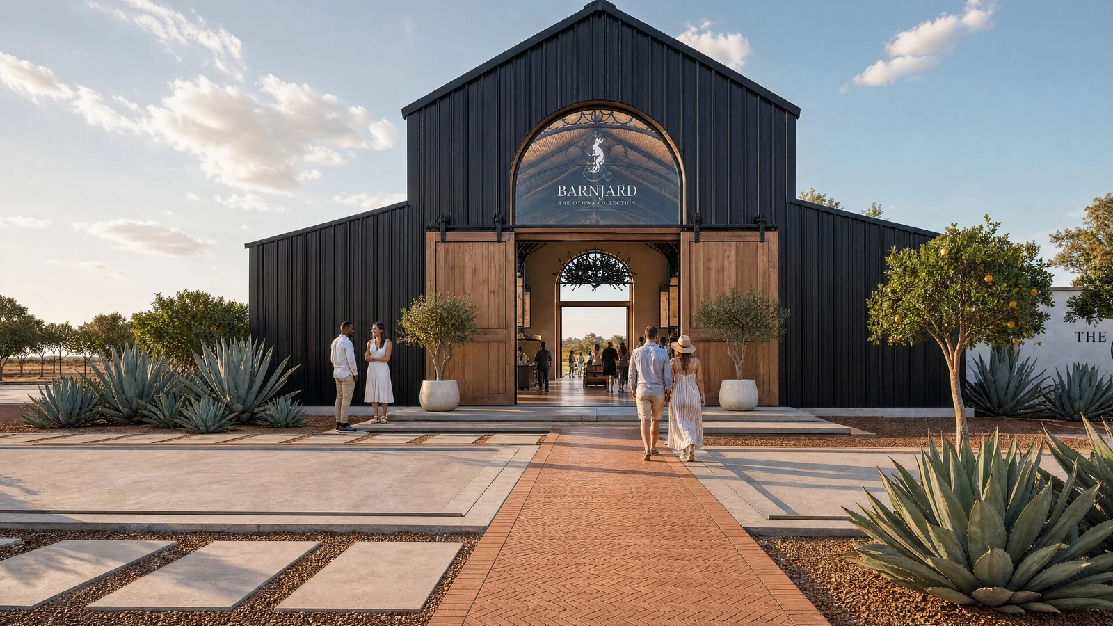 A tall black barn with an arched glazed entrance and open timber doors at the end of a herringbone brick path, visitors walking in